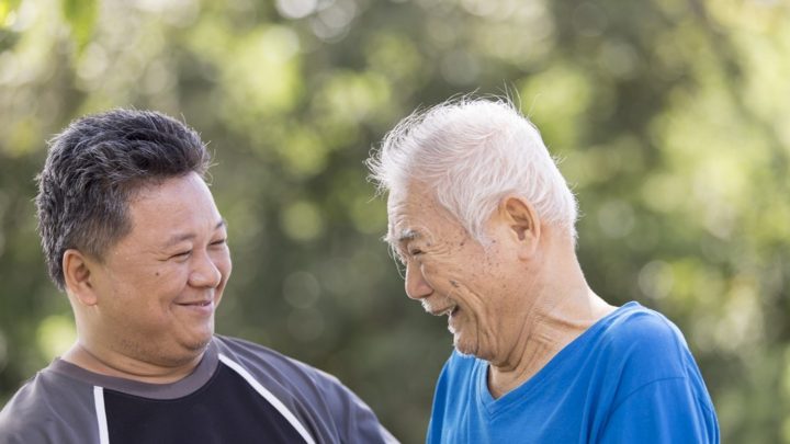 An adult son and older adult father, who has Alzheimer's disease, outdoors smiling at each other.