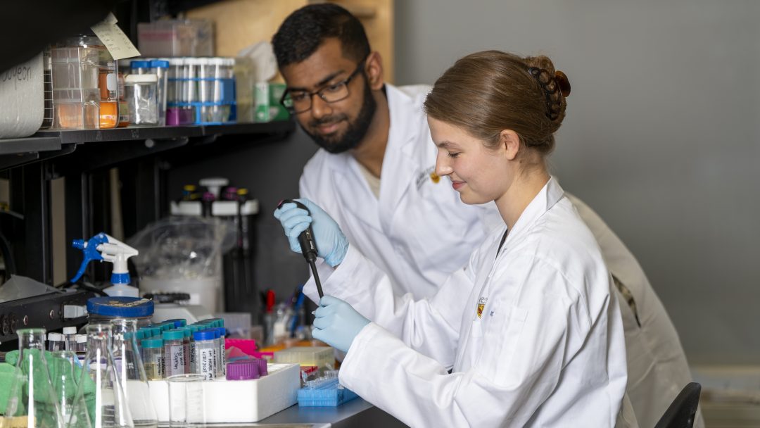 Two students in lab coats working at a lab table.