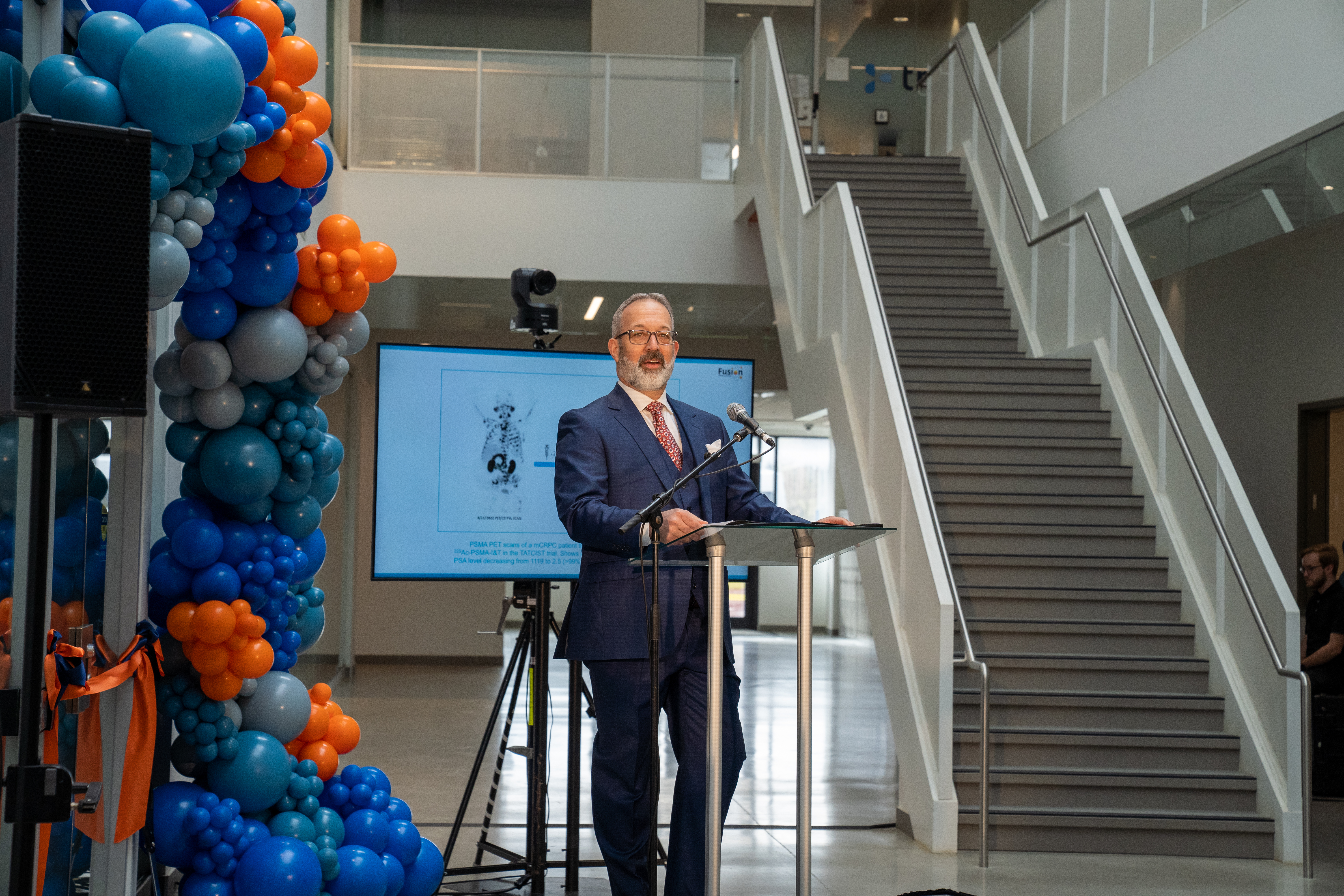 CEO John Valliant stands in front of a tv and multicoloured balloons, giving a presentation.